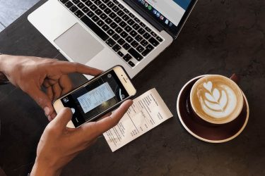 Image of a hand holding an Iphone taking a photo of a receipt, closely to his coffee and laptop.